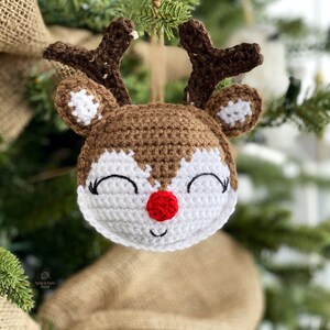 Puede incluir: Un adorno de reno de ganchillo con cuernos marrones, una cara blanca y una nariz roja. El adorno est&aacute; colgado de un &aacute;rbol de Navidad.