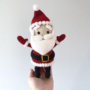 Puede incluir: Un mu&ntilde;eco de Pap&aacute; Noel de crochet con barba blanca, gorro rojo y cintur&oacute;n negro. El mu&ntilde;eco tiene los brazos extendidos y una expresi&oacute;n amigable.