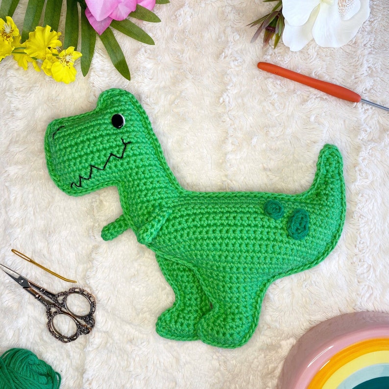 Pode incluir: Brinquedo de dinossauro de croch&ecirc; verde com olhos pretos e um sorriso costurado. O dinossauro tem dois c&iacute;rculos verdes nas costas.