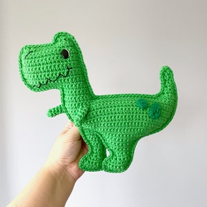 Pode incluir: Brinquedo de dinossauro de croch&ecirc; verde com olhos pretos e uma boca sorridente. O dinossauro tem manchas verdes nas costas.