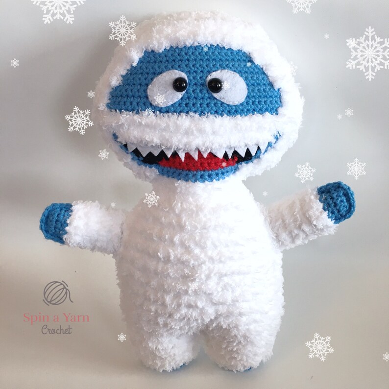 Puede incluir: Un mu&ntilde;eco de nieve blanco de crochet con detalles azules y una boca roja. El mu&ntilde;eco de nieve tiene ojos negros y una textura blanca y esponjosa. El mu&ntilde;eco de nieve est&aacute; de pie sobre un fondo blanco con copos de nieve.