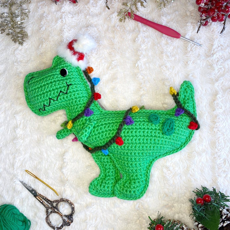 Pode incluir: Um dinossauro de croch&ecirc; verde usando um chap&eacute;u de Papai Noel vermelho e branco com luzes coloridas enroladas ao redor. O dinossauro est&aacute; sentado em uma superf&iacute;cie branca e fofa.