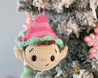 May include: A crocheted elf ornament with a pink and green hat and a brown face. The elf is hanging on a snowy Christmas tree.