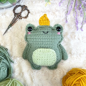 May include: A green crocheted frog wearing a yellow crocheted crown. The frog has black eyes and pink cheeks.