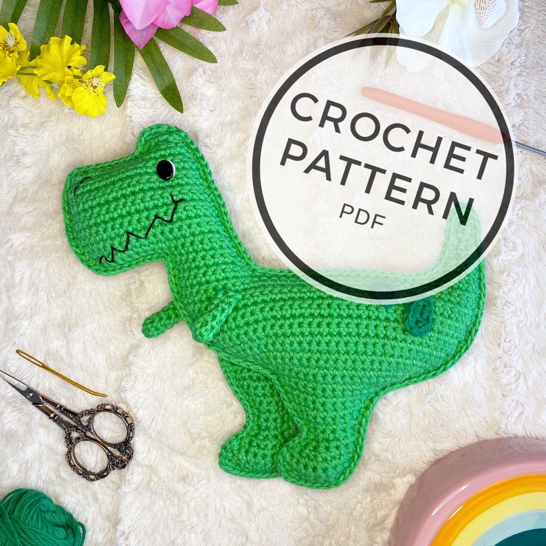 Pode incluir: Brinquedo de dinossauro de croch&ecirc; verde com olhos pretos e um sorriso costurado. O brinquedo est&aacute; sobre uma superf&iacute;cie branca e fofa com uma flor rosa e branca no fundo. Um c&iacute;rculo com o texto "CROCHET PATTERN PDF" est&aacute; no canto superior direito da imagem.