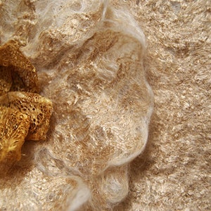 K&ouml;nnte beinhalten: Eine goldfarbene Stoffblume mit einem Spitzenmuster und klaren Strasssteinen liegt auf einem strukturierten, beigefarbenen Hintergrund.