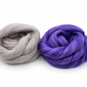 May include: Two bundles of wool roving. One is a light gray color, and the other is a vibrant purple with lighter purple streaks. The wool is tightly coiled, suggesting it is ready for crafting projects like felting or spinning.