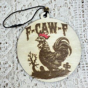 May include: A round wooden ornament with the text "F-CAW-F" at the top. The ornament features a detailed illustration of a rooster with a skull-like head, a red comb, and a large tail. A small tree and a tombstone are also depicted.