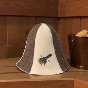 May include: A sauna hat with a rounded top and a wide brim. The hat is primarily white with brown side panels and a stitched edge. A small embroidered design of green leaves and black stems is on the front. The hat sits on a wooden bench.
