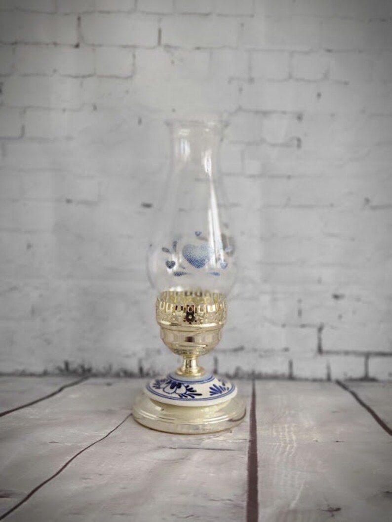 Op de afbeelding: Een vintage olielamp met een heldere glazen schoorsteen en een goudkleurige metalen voet. De voet heeft blauwe bloemmotieven op een witte achtergrond. De lamp staat tegen een witte bakstenen muur.