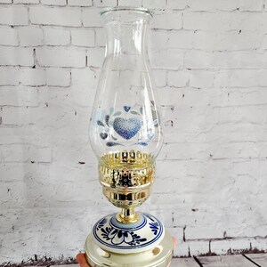Op de afbeelding: Glazen olielamp met een goudkleurige metalen voet en een decoratief blauw en wit bloemmotief. Het glazen gedeelte heeft een blauw hart- en bloemmotief. De lamp wordt in een hand gehouden.