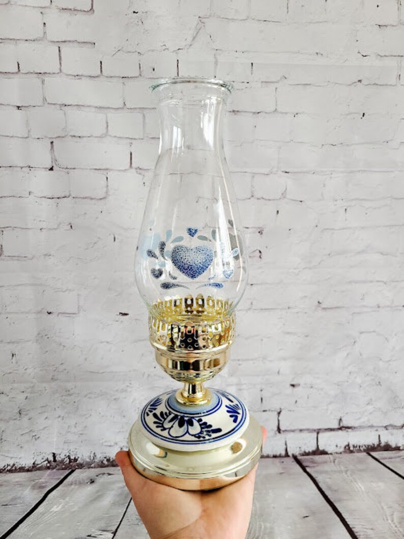 Op de afbeelding: Glazen olielamp met een goudkleurige metalen voet en een decoratief blauw en wit bloemmotief. Het glazen gedeelte heeft een blauw hart- en bloemmotief. De lamp wordt in een hand gehouden.