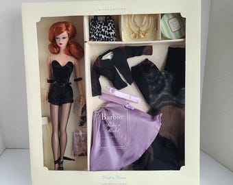Puede incluir: Un conjunto de Barbie Fashion Model Collection en caja. La muñeca tiene el pelo rojo y lleva un atuendo negro. Los accesorios incluyen un bolso con estampado de leopardo, joyas y una capa morada. La caja dice "Edición Limitada" y "Dusk to Dawn".