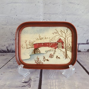 May include: A framed embroidered picture of a red covered bridge in a snowy winter scene. The bridge is over a stream with trees and bushes on either side. There are small animals in the snow.
