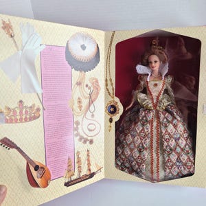 Puede incluir: Muñeca de una figura histórica en una caja. La muñeca lleva un vestido detallado con una falda estampada y un cuello blanco. La caja incluye ilustraciones de accesorios y un barco.