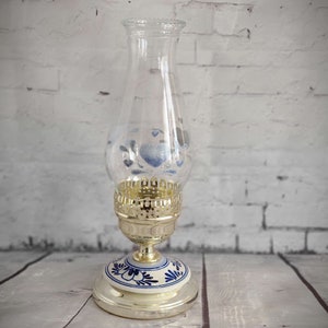 Op de afbeelding: Een heldere glazen olielamp met een goudkleurige metalen voet en een wit en blauw bloemmotief op de voet. De glazen schoorsteen heeft blauwe bloemaccenten. De lamp is ontworpen voor decoratief gebruik.