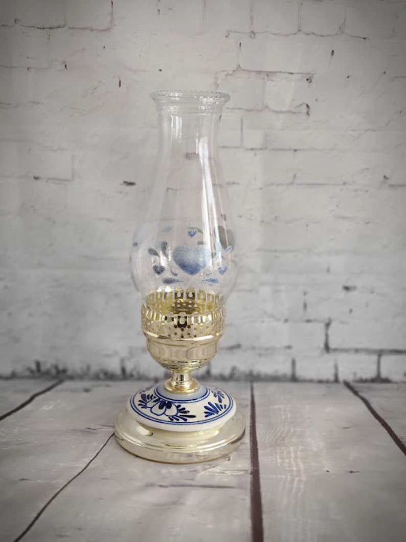 Op de afbeelding: Een heldere glazen olielamp met een goudkleurige metalen voet en een wit en blauw bloemmotief op de voet. De glazen schoorsteen heeft blauwe bloemaccenten. De lamp is ontworpen voor decoratief gebruik.