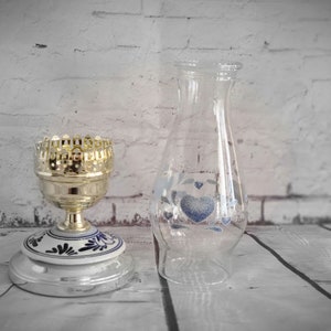 Op de afbeelding: Een heldere glazen vaas met blauwe hart- en bloemmotieven naast een goudkleurige metalen kandelaar met een wit en blauw keramisch voetstuk. De kandelaar heeft een decoratieve, opengewerkte bovenkant.