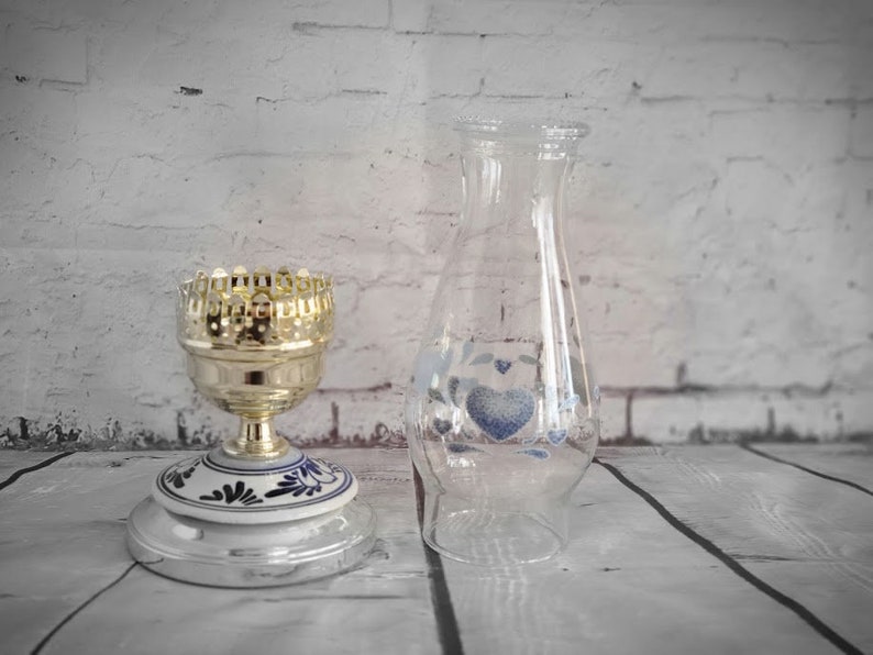 Op de afbeelding: Een heldere glazen vaas met blauwe hart- en bloemmotieven naast een goudkleurige metalen kandelaar met een wit en blauw keramisch voetstuk. De kandelaar heeft een decoratieve, opengewerkte bovenkant.