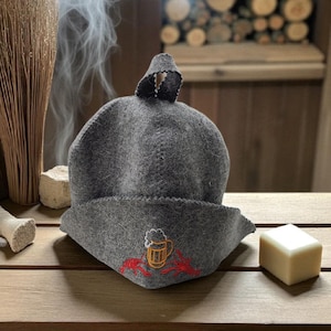 May include: A grey felt sauna hat with a beer mug and lobster embroidery. The hat has a rounded crown and a wide brim. The background includes a wooden surface and a wooden wall.