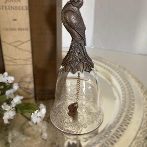 Op de afbeelding: Een decoratieve glazen bel met een gedetailleerd zilverkleurig vogelbeeldje erop. De bel heeft een geëtst bloemmotief en een kleine klepel. De bel wordt gepresenteerd op een decoratief zilverkleurig dienblad, met boeken op de achtergrond.