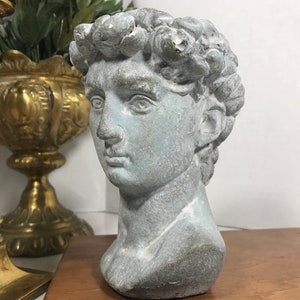 Può includere: Busto grigio-blu di una figura maschile classica con capelli ricci. La scultura ha un aspetto invecchiato, che suggerisce un'estetica antica o antica. Il busto è esposto su una superficie di legno, con un candelabro in ottone e un vaso decorativo sullo sfondo.