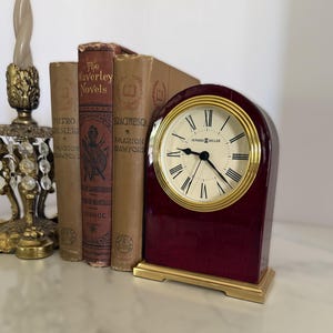 May include: A Howard Miller mantel clock with a deep burgundy, high-gloss finish and a gold-tone metal base. The clock face is cream-colored with black Roman numerals and black hands, featuring the Howard Miller logo in the center.