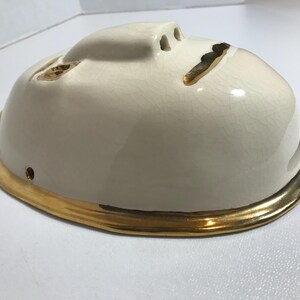 May include: White ceramic mask with a gold rim. The mask has a simple face design with three holes for the eyes and nose.