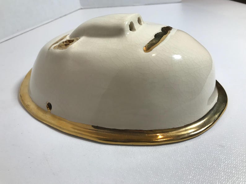May include: White ceramic mask with a gold rim. The mask has a simple face design with three holes for the eyes and nose.