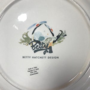 May include: White ceramic plate featuring a blue and green bird illustration with the text "Betty" in a stylised font. The plate also includes the text "Betty Hatchett Design" and "Made in China, Fabriqu&eacute; en Chine, Hand wash recommended".