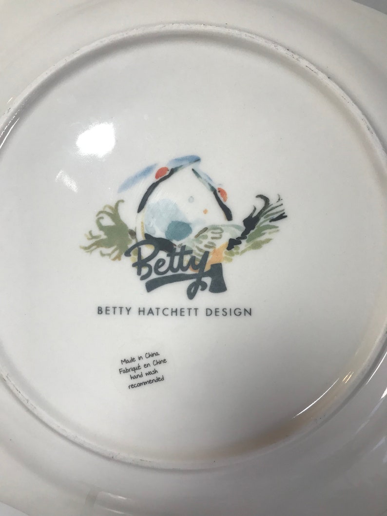 May include: White ceramic plate featuring a blue and green bird illustration with the text "Betty" in a stylised font. The plate also includes the text "Betty Hatchett Design" and "Made in China, Fabriqu&eacute; en Chine, Hand wash recommended".
