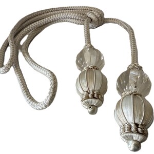 May include: A pair of decorative curtain tiebacks. The tiebacks feature a light beige rope with a knot at the top. Each tieback has a clear, round bead above a ribbed, light beige, spherical tassel. The tassels have a small, silver-colored metal end.