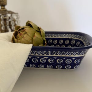 Puede incluir: Una fuente rectangular de cerámica con un patrón de espirales azul oscuro y blanco en los lados y el borde interior. Una alcachofa verde descansa dentro de la fuente, parcialmente cubierta por una servilleta de tela color hueso.