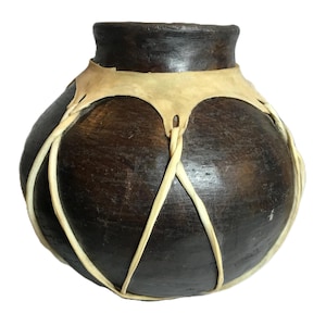 Könnte beinhalten: Ein handgefertigter, dunkelbrauner Keramiktopf mit breitem Körper und schmalem Hals. Der Topf ist mit einem hellbraunen Lederband und cremefarbenen gewebten Akzenten verziert. Der Topf hat eine rustikale, erdige Ästhetik.