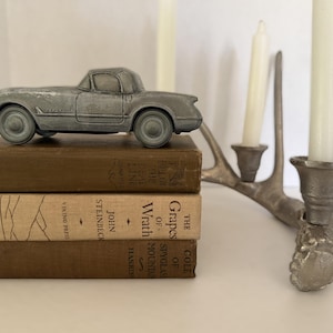 May include: A gray die-cast model car sits atop a stack of vintage books. The books are various shades of brown and tan. A silver-colored antler-style candle holder with white candles is in the background.