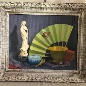 May include: A framed oil painting of a still life featuring a white porcelain figurine, a green folding fan, a gold bowl, a blue and white bowl, and a wooden spoon on a purple and red patterned cloth.