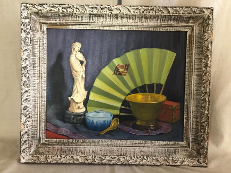May include: A framed oil painting of a still life featuring a white porcelain figurine, a green folding fan, a gold bowl, a blue and white bowl, and a wooden spoon on a purple and red patterned cloth.