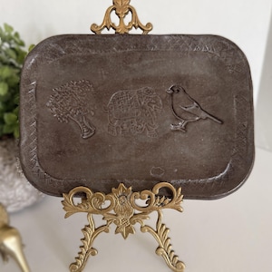 May include: A dark grey rectangular tray featuring embossed images of a tree, elephant, and bird. The tray is supported by a gold-coloured metal stand with ornate floral and leaf designs. The tray's border has a decorative pattern.