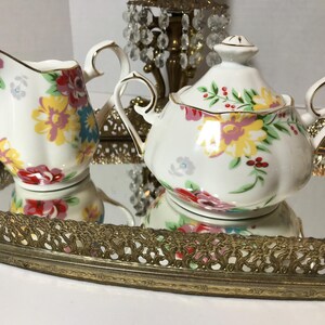 May include: A white porcelain creamer and sugar bowl set with a floral pattern in shades of pink, yellow, and red. The set is displayed on a gold metal tray with a decorative design.