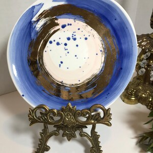May include: A white ceramic plate with a blue and gold abstract design. The plate has a pink and white center with gold brushstrokes and blue speckles. The plate is on a gold metal stand.