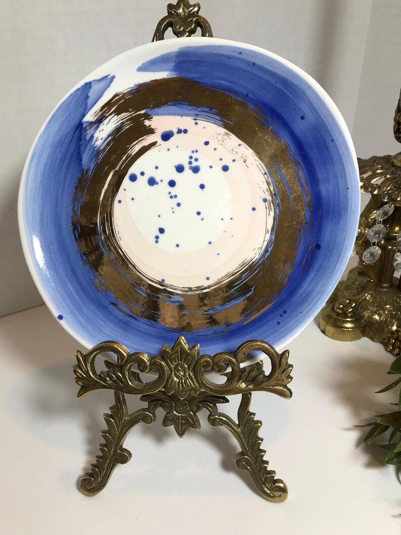 May include: A white ceramic plate with a blue and gold abstract design. The plate has a pink and white center with gold brushstrokes and blue speckles. The plate is on a gold metal stand.
