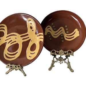 May include: Two brown ceramic plates with gold abstract designs. The plates are displayed on decorative brass stands. One plate features a large, stylized design, while the other has a wavy pattern with dots.