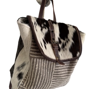 May include: A backpack with a cowhide flap and accents. The flap features a brown and white pattern, with a brown leather strap and buckle. The body of the bag is a mix of striped and solid fabric in shades of brown and beige.