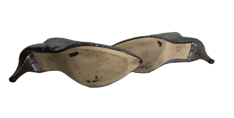 May include: A pair of silver sequined high-heeled shoes. The shoes have a pointed toe and a stiletto heel. The soles are beige with some wear. The shoes are angled, showing the underside and side profile.