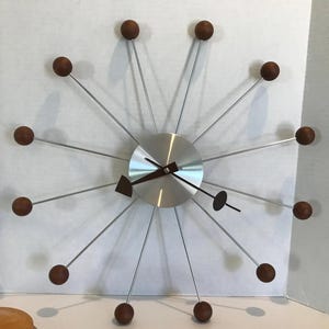 Puede incluir: Un reloj de pared moderno de mediados de siglo con una esfera de metal cepillado y detalles de madera marrón. El reloj presenta largas varillas plateadas que se extienden desde el centro, cada una terminada en una esfera marrón. Las manecillas son marrones y negras.