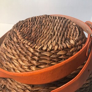 Op de afbeelding: Een geweven mand met een bruine leren riem eromheen gewikkeld. De riem is oranje en heeft een gesp.