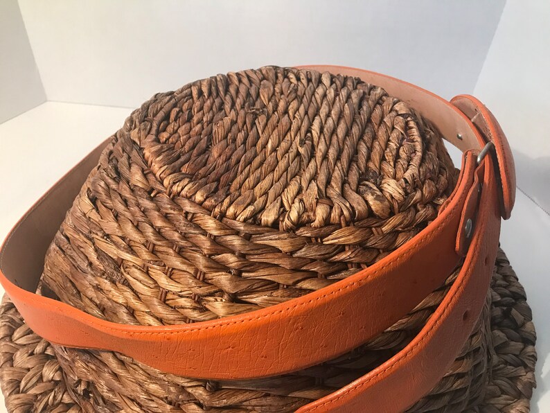 Op de afbeelding: Een geweven mand met een bruine leren riem eromheen gewikkeld. De riem is oranje en heeft een gesp.