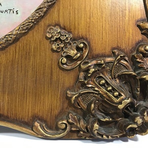 May include: Close-up of a wooden frame with ornate gold accents. The frame has a carved design and a distressed finish. The text "Gloria Curtis" is written in black ink on the top left corner of the frame.