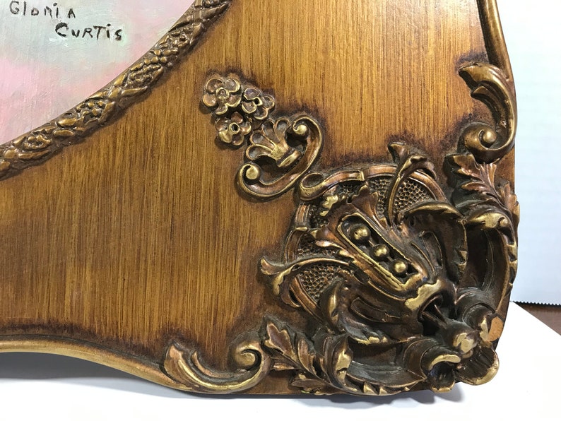 May include: Close-up of a wooden frame with ornate gold accents. The frame has a carved design and a distressed finish. The text "Gloria Curtis" is written in black ink on the top left corner of the frame.
