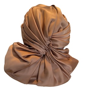 Puede incluir: Un turbante drapeado en un cálido color marrón dorado. La tela es lisa y parece ser satén o un material similar, con múltiples pliegues y fruncidos que crean una forma voluminosa. El turbante está diseñado para ser usado en la cabeza.